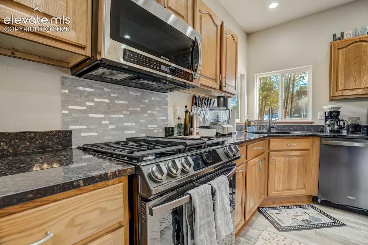 Kitchen featuring a stainless steel range with overhead microwave, dark granite-finish countertops, wood cabinetry, and a mosaic tile backsplash