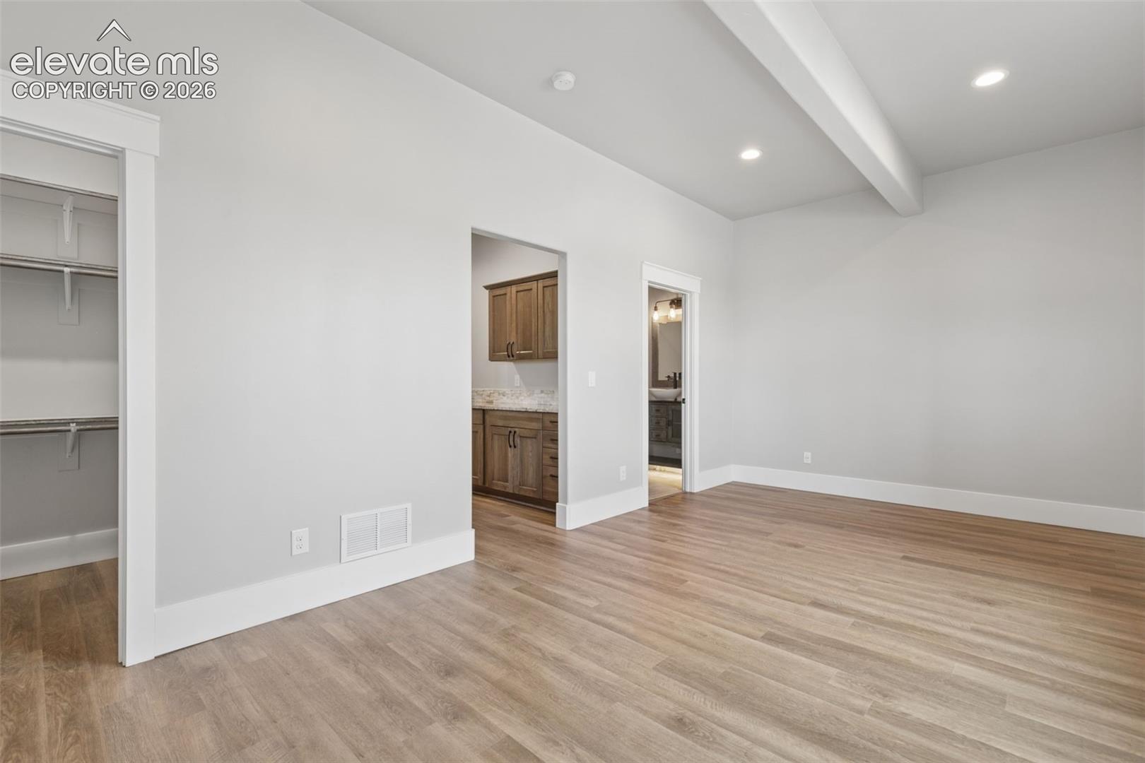 Vaulted ceiling with exposed beam, recessed lighting, wood-finish flooring, built-in closet shelving, and a secondary room featuring wood cabinetry and a stone-finish backsplash
