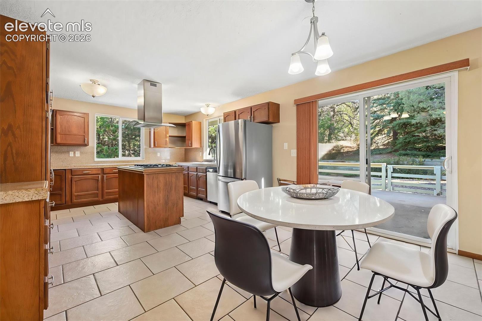 Kitchen featuring wood cabinetry, stainless steel island-mounted range hood, stainless steel refrigerator, stone-finish countertops, and tile flooring