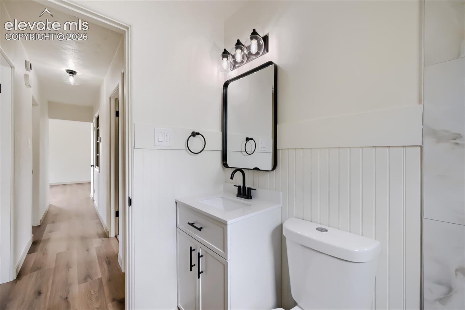 Vanity with integrated sink, black-framed mirror, three-light vanity fixture, beadboard wainscoting, and wood-finish flooring
