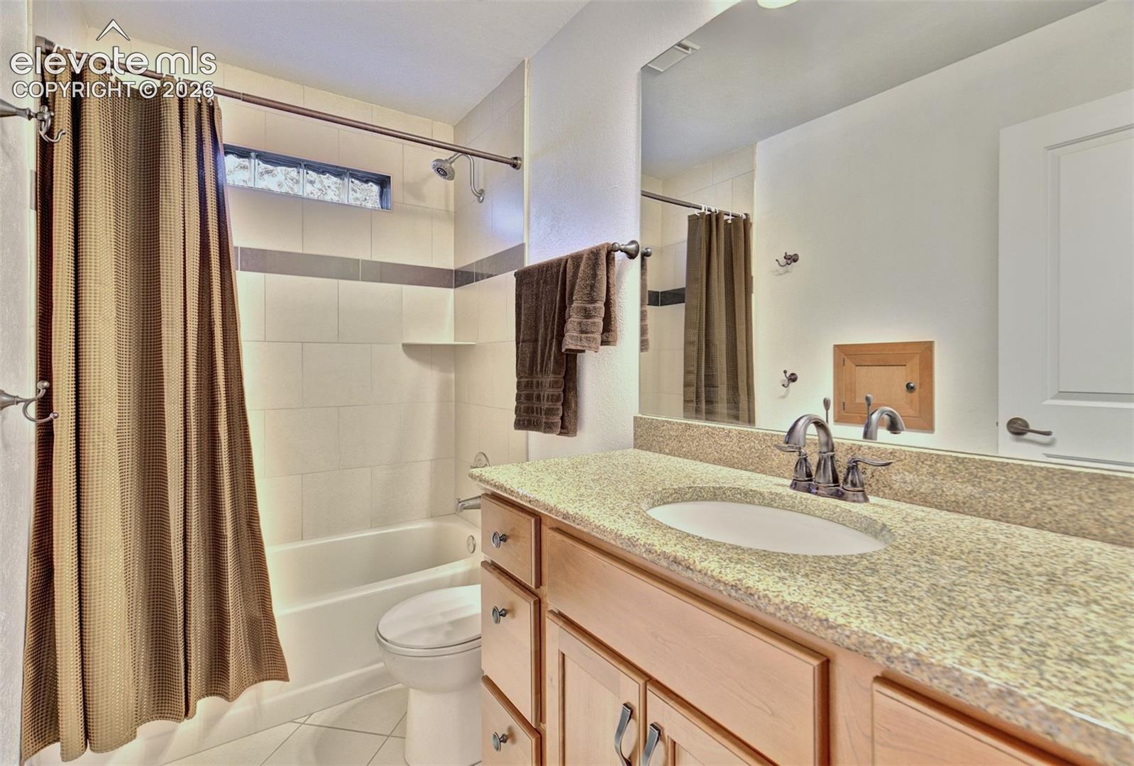 Bathroom featuring a vanity with stone-finish countertop, undermount sink, and multi-drawer storage, alongside a tub-shower combination with light-toned wall tile, a horizontal glass block window, and a large wall-mounted mirror