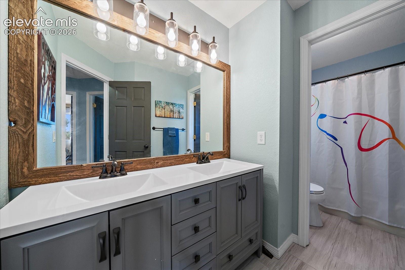 Dual-sink vanity with gray shaker-style cabinetry, white countertop, integrated rectangular basins, and matte black hardware, accented by a large custom wood-framed mirror and multi-bulb industrial lighting