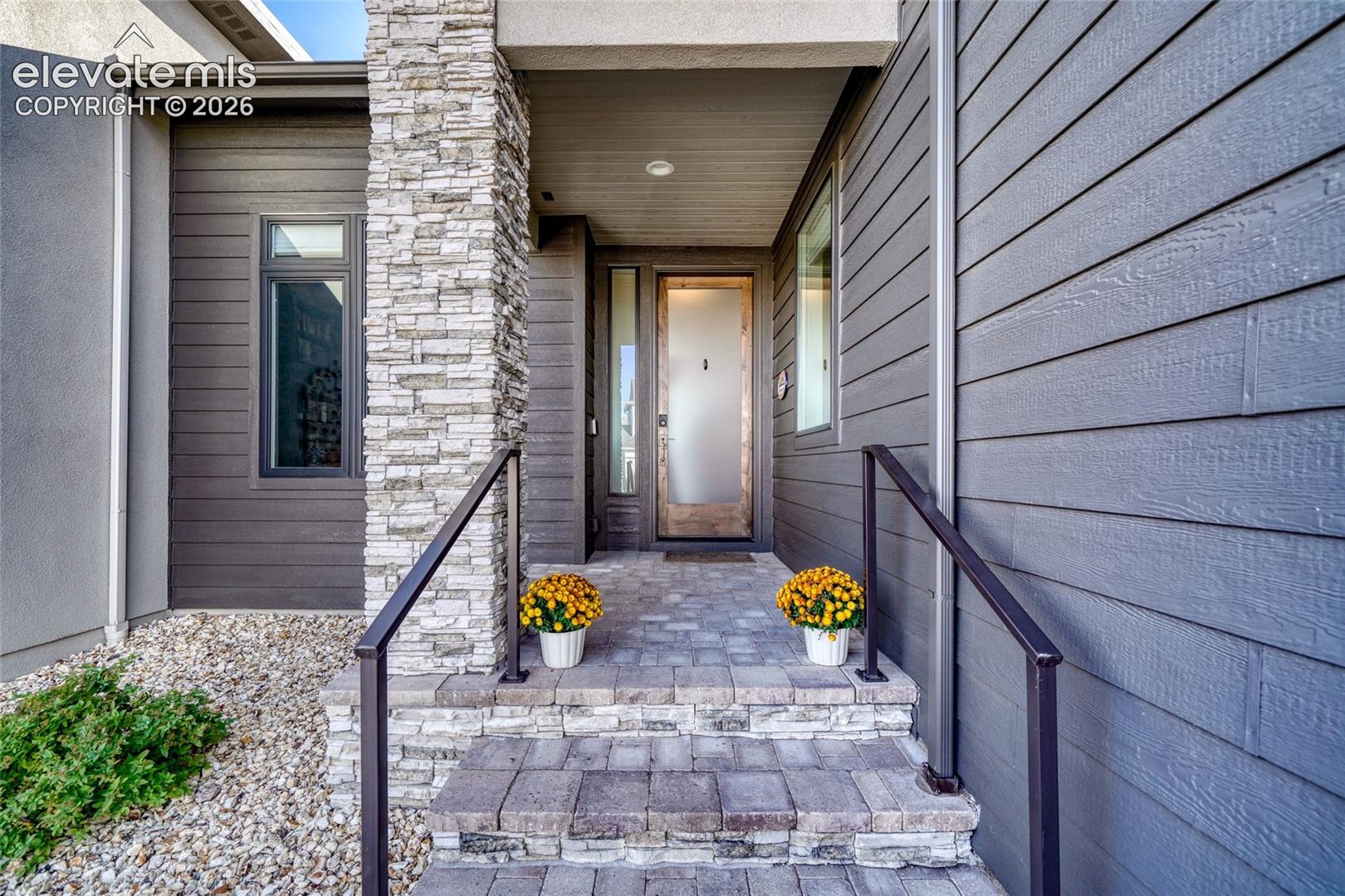Covered entryway featuring horizontal lap siding, a natural stacked stone pillar, paver-style steps, a frosted glass entry door with a vertical sidelight, and metal handrails