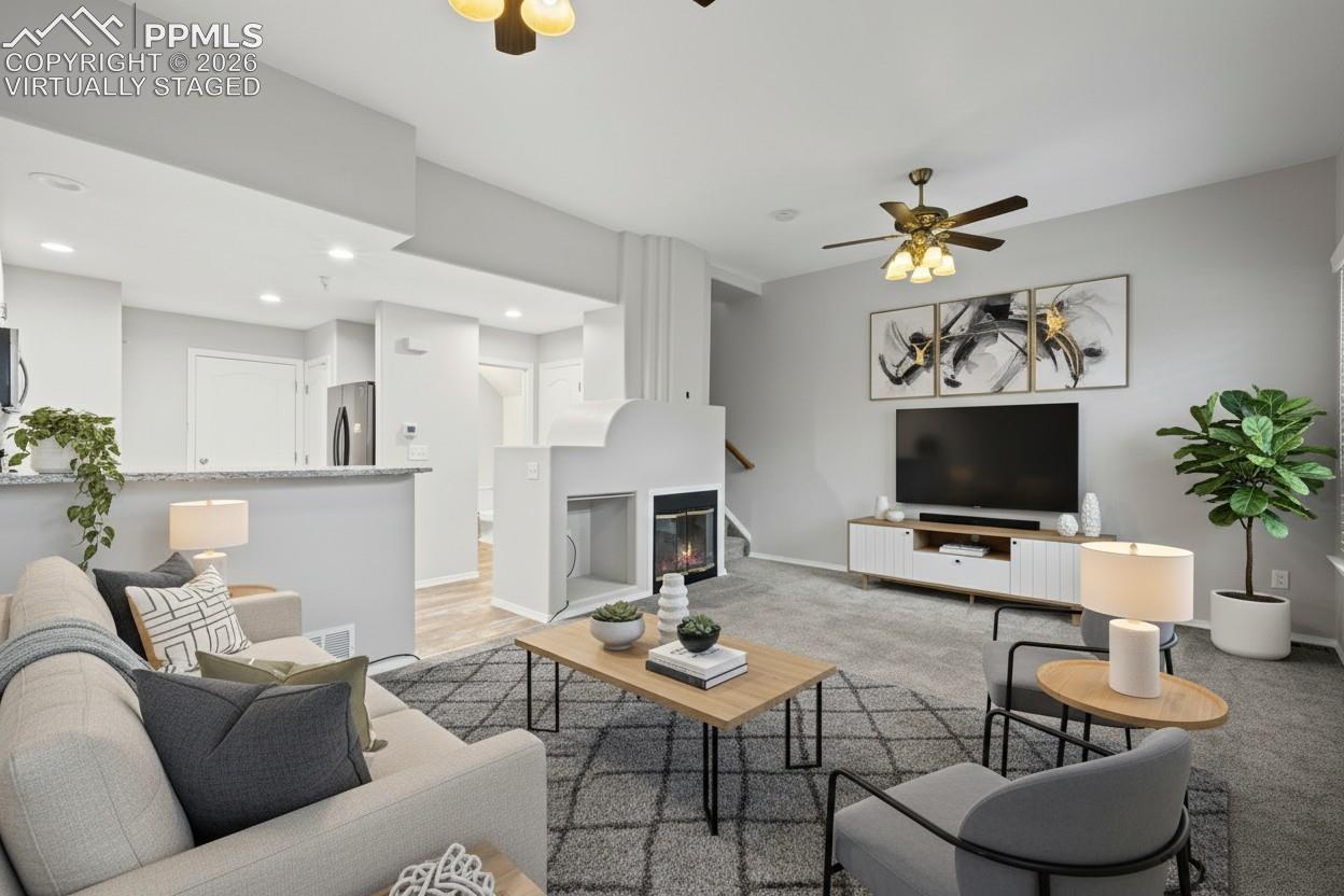 Living room featuring custom window shutters, a ceiling fan, carpet flooring, stairway, and a glass covered fireplace. This photo is virtually staged
.