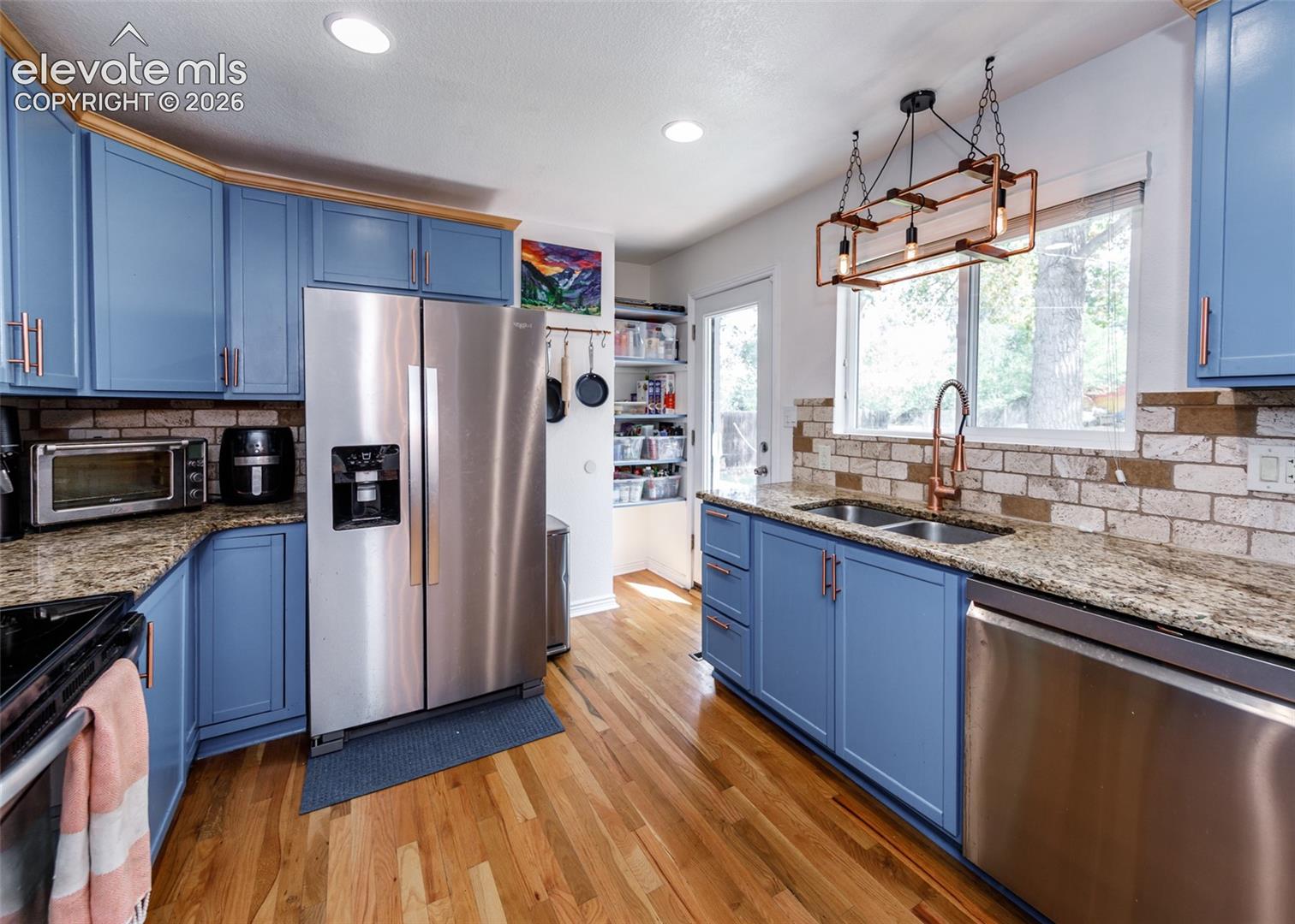 Hardwood flooring, light blue cabinetry with rose gold hardware, granite countertops, brick-style backsplash, and stainless steel appliances