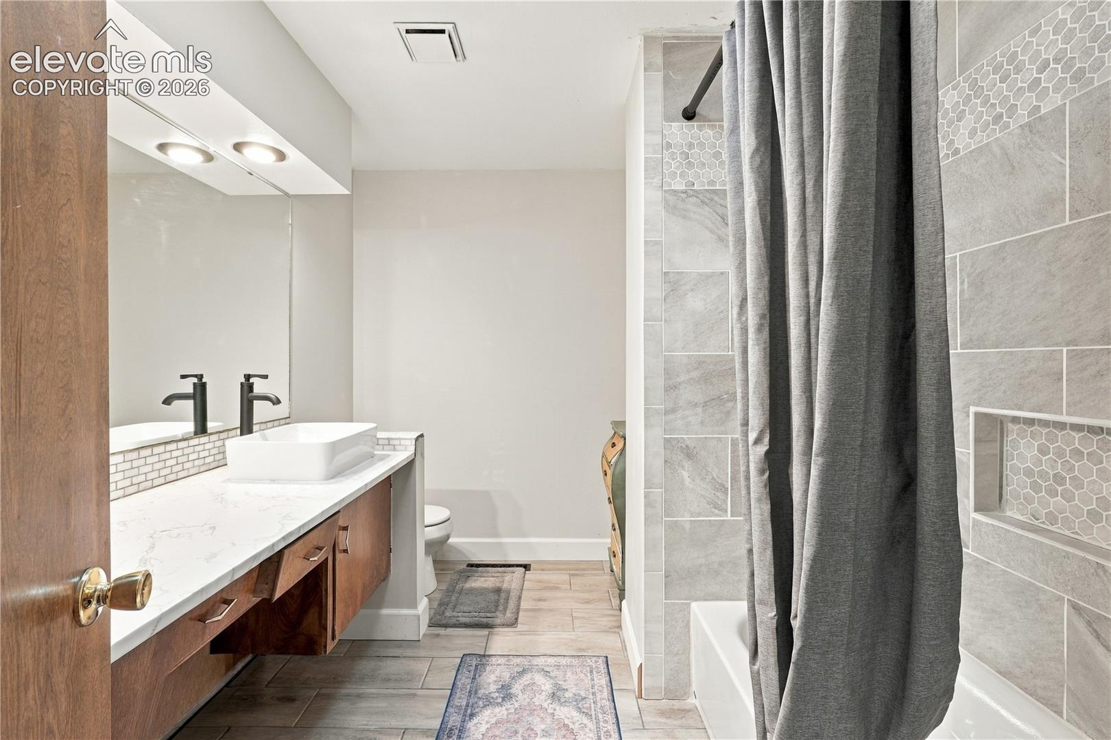 Bathroom featuring wood-finish cabinetry, white countertop with a rectangular vessel sink, matte black fixtures, wood-finish tile flooring, and a tiled shower surround with a built-in recessed niche