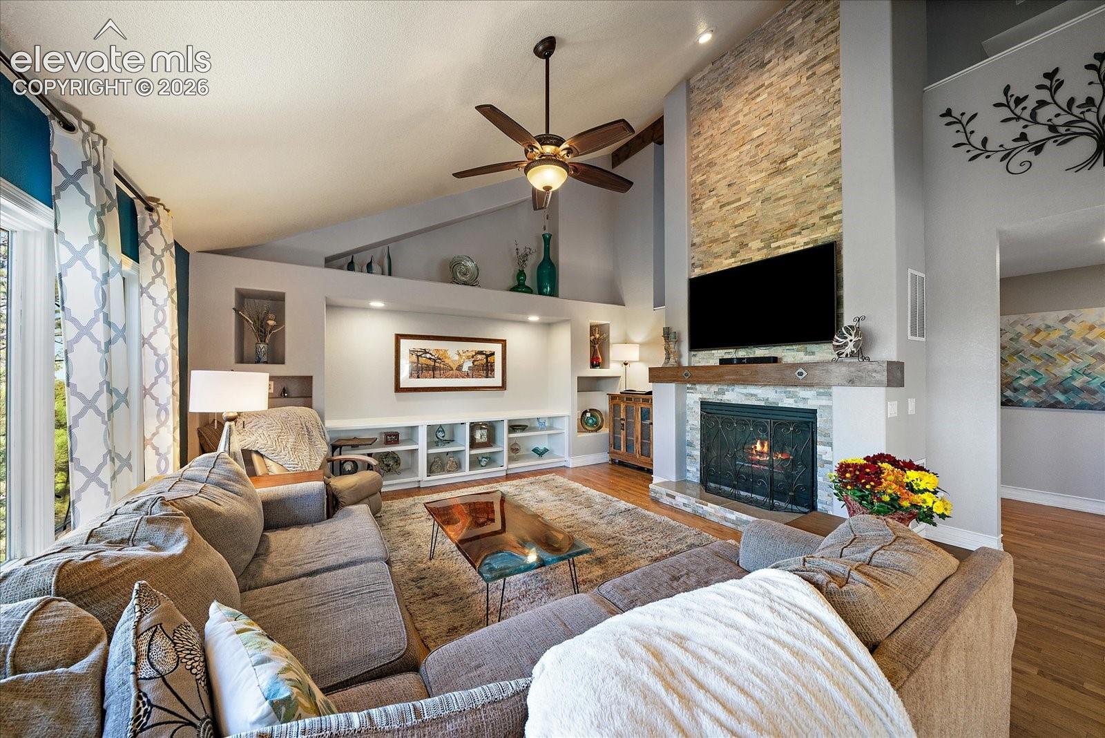 Living area featuring a vaulted ceiling, floor-to-ceiling stacked stone fireplace, custom built-in cabinetry, recessed shelving, and wood-finish flooring