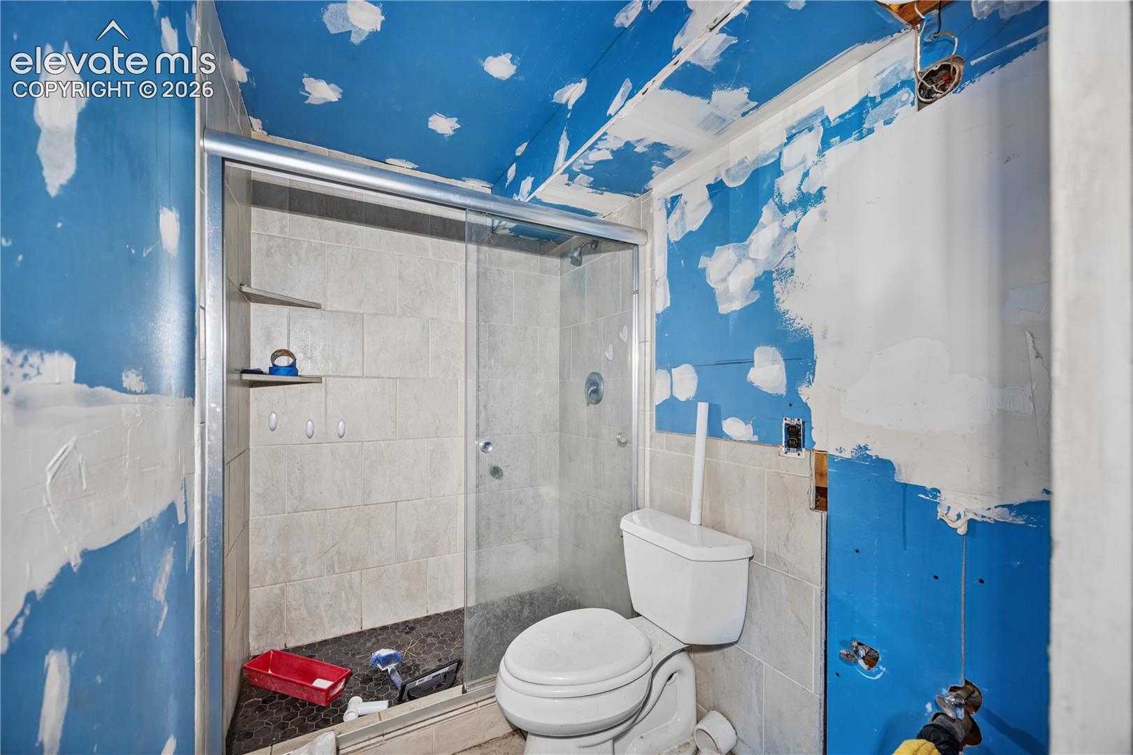 Shower enclosure with sliding glass door, tiled wall surround, stone-tiled floor, and dual built-in corner shelving, featuring an installed porcelain toilet
