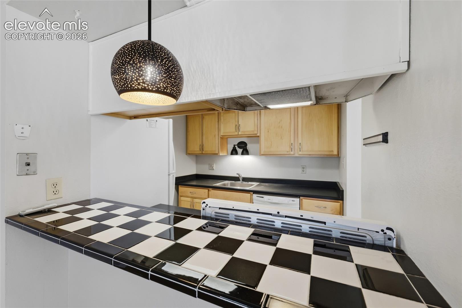 Kitchen featuring a checkerboard-patterned tile breakfast bar, natural-finish wood cabinetry, a dark solid-surface countertop, and a metal pendant light fixture