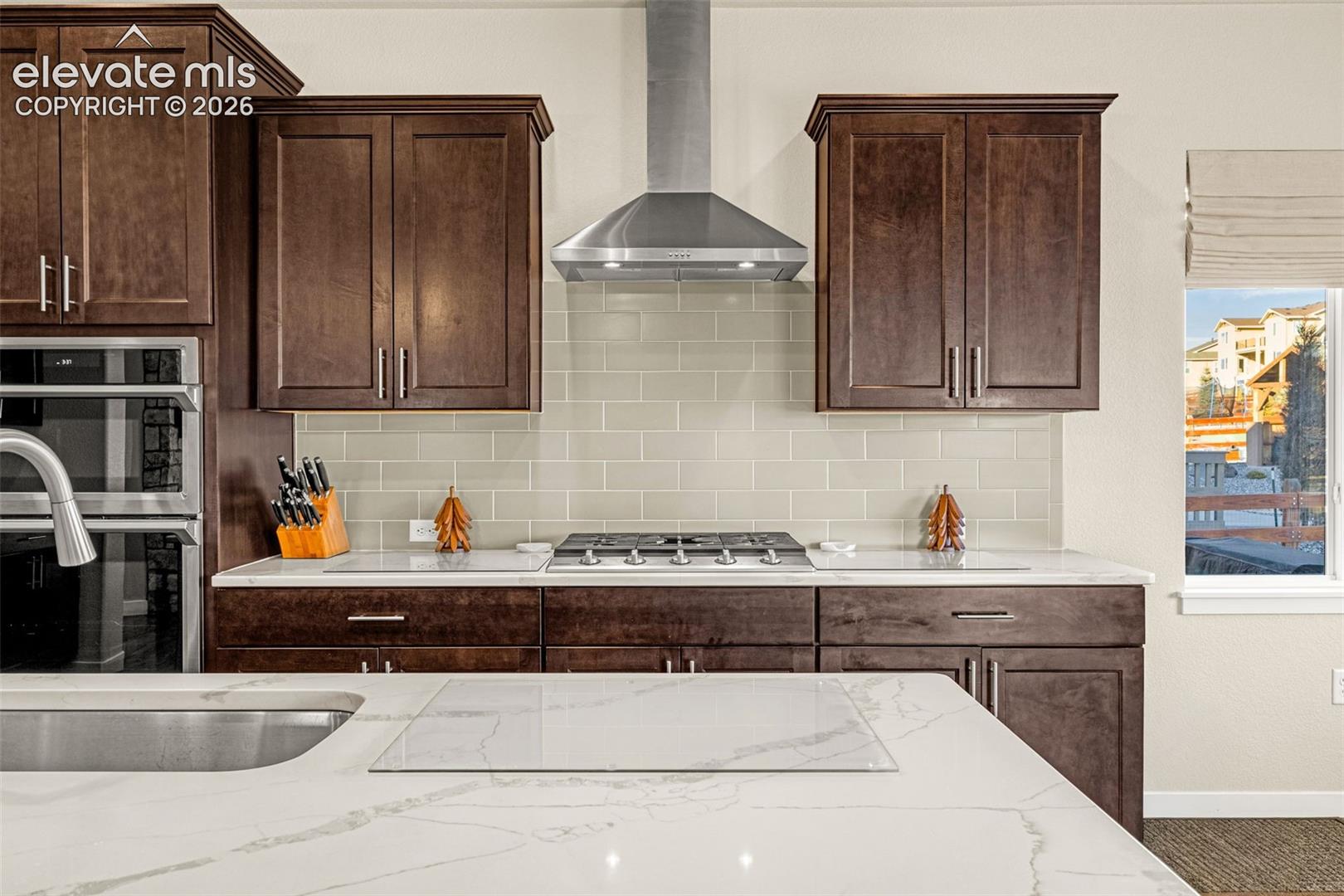 Kitchen featuring dark wood cabinetry, light-toned stone-finish countertops, subway tile backsplash, stainless steel gas cooktop with chimney-style range hood, and built-in double wall oven