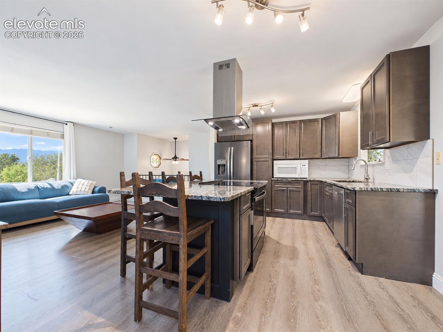 Updated kitchen with dark cabinetry, granite-style countertops, stainless steel appliances, a large center island with seating, and a contemporary island range hood. The open layout connects naturally with the adjoining living area, where mountain views are visible through the window.