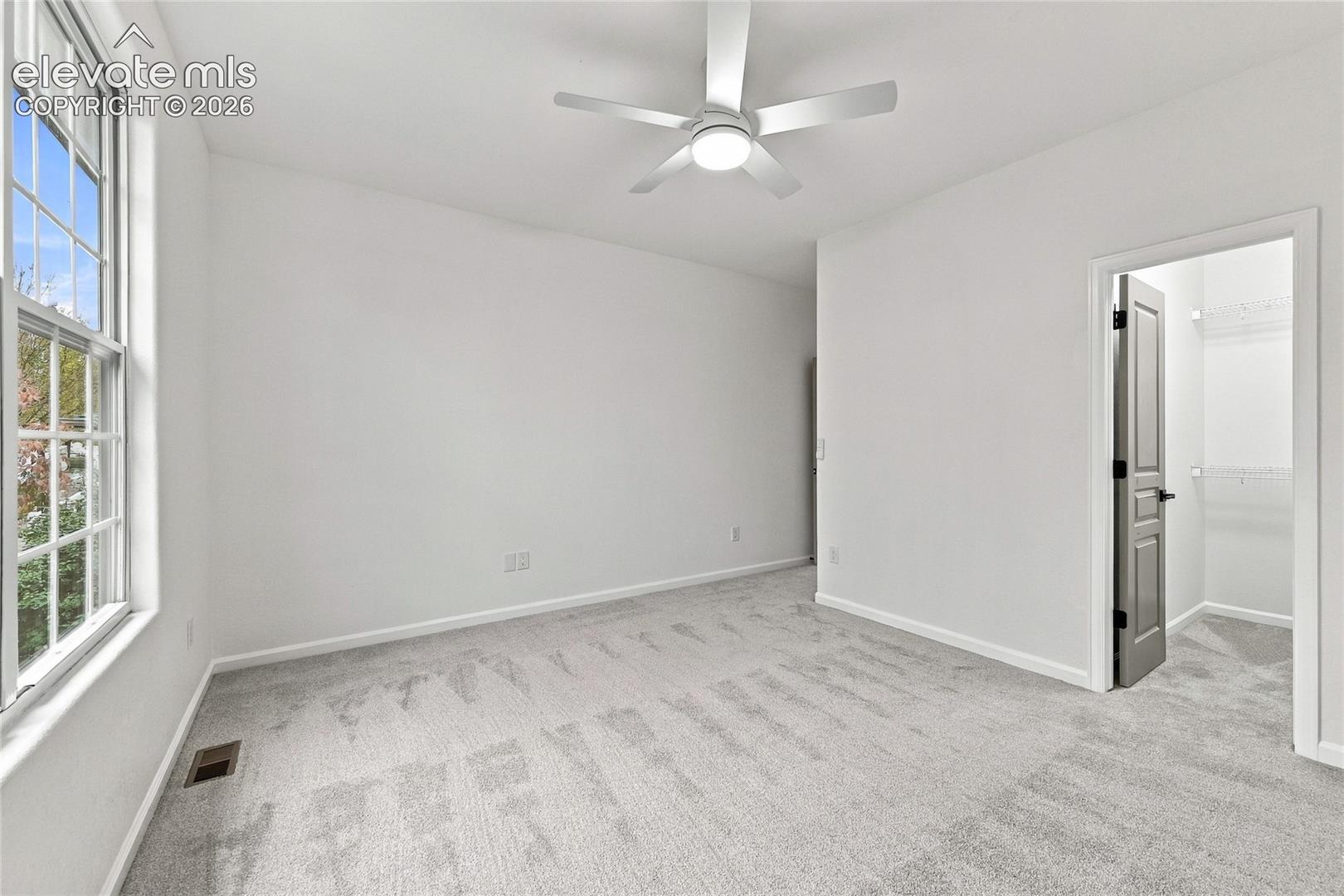 Neutral-toned carpeting, multi-pane window, white ceiling fan with integrated lighting, and interior door opening to a closet with wire shelving