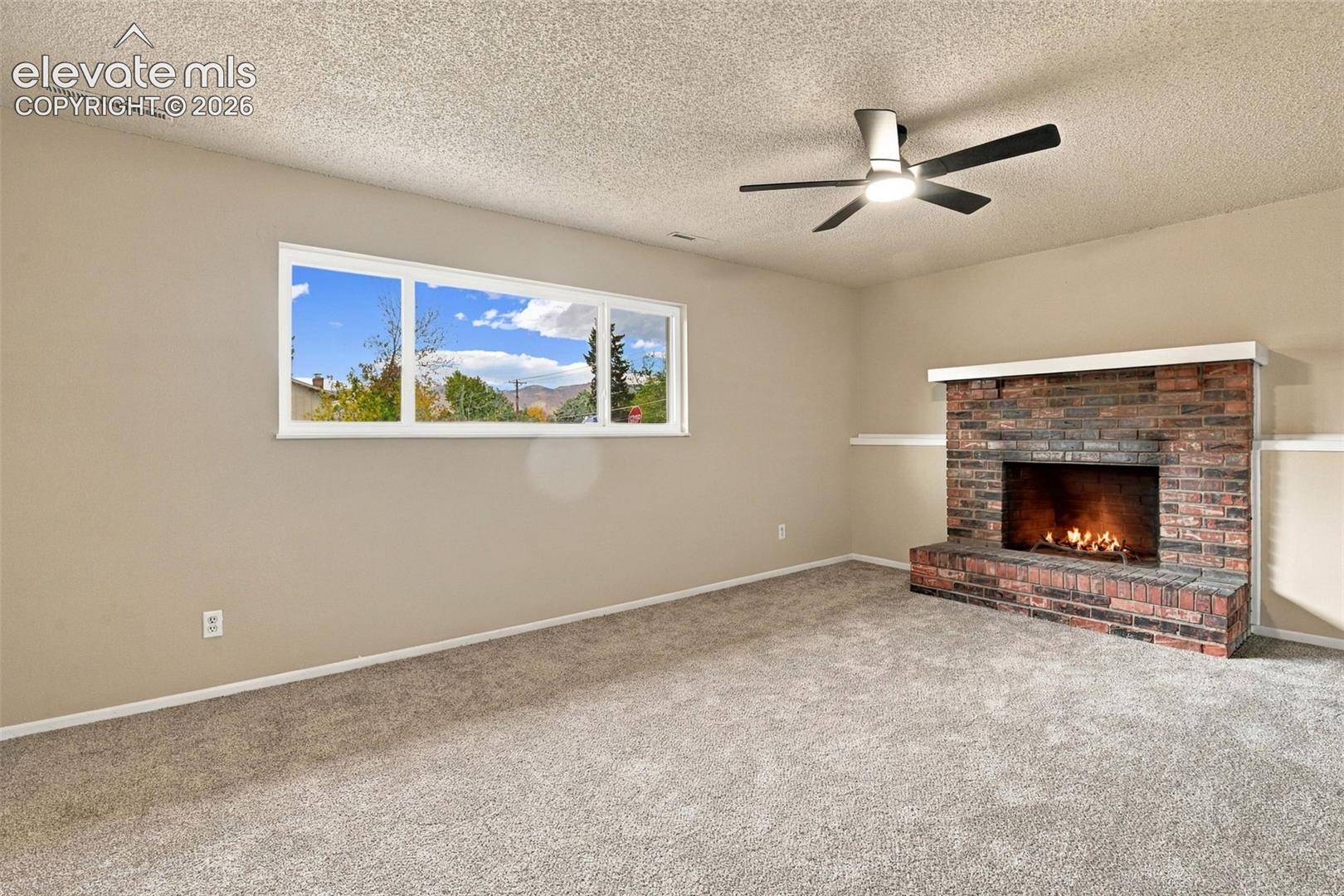 Room featuring a brick fireplace with a mantle, neutral wall-to-wall carpeting, a large picture window, and a ceiling fan