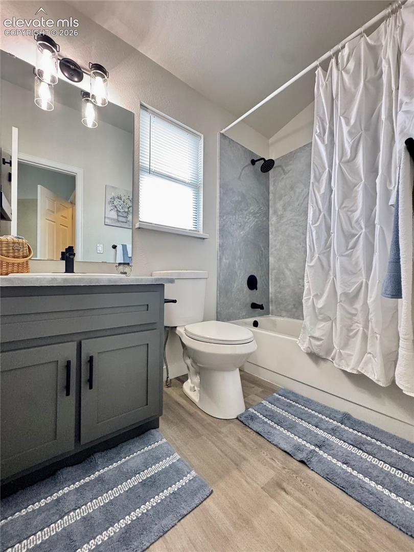 Bathroom featuring a grey shaker-style vanity, matte black hardware, wood-finish flooring, a wall-mounted three-light fixture, and a tub-shower surround with a stone-pattern finish