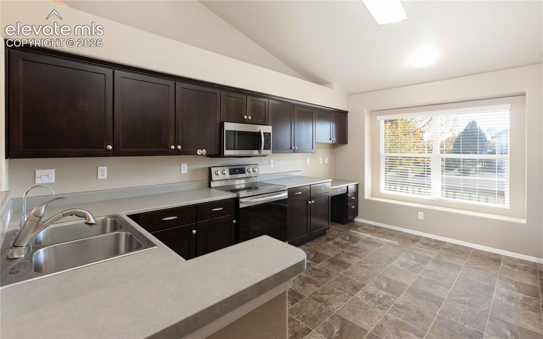 Kitchen featuring dark wood cabinetry, solid surface countertops, stainless steel appliances, tile flooring, and a large window