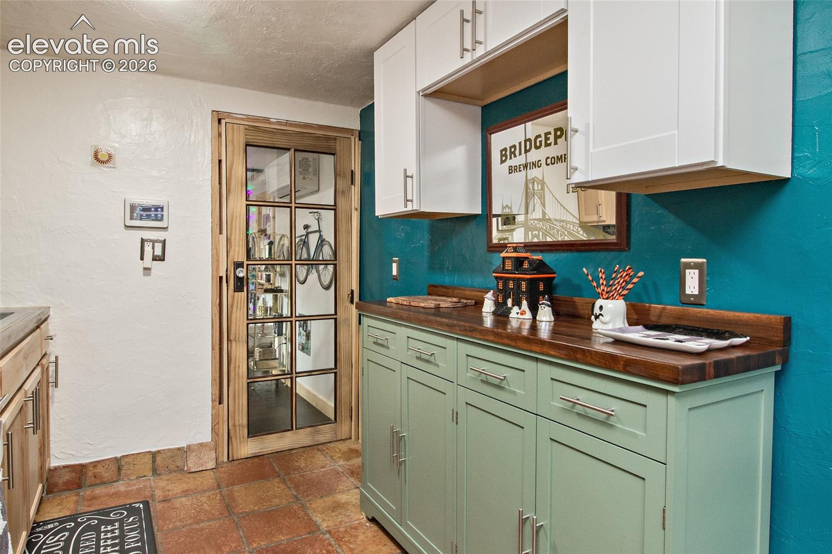Built-in cabinetry featuring white upper cabinets and sage green lower base cabinets, wood-finish countertop, terracotta tile flooring, and a glass-paneled wooden door