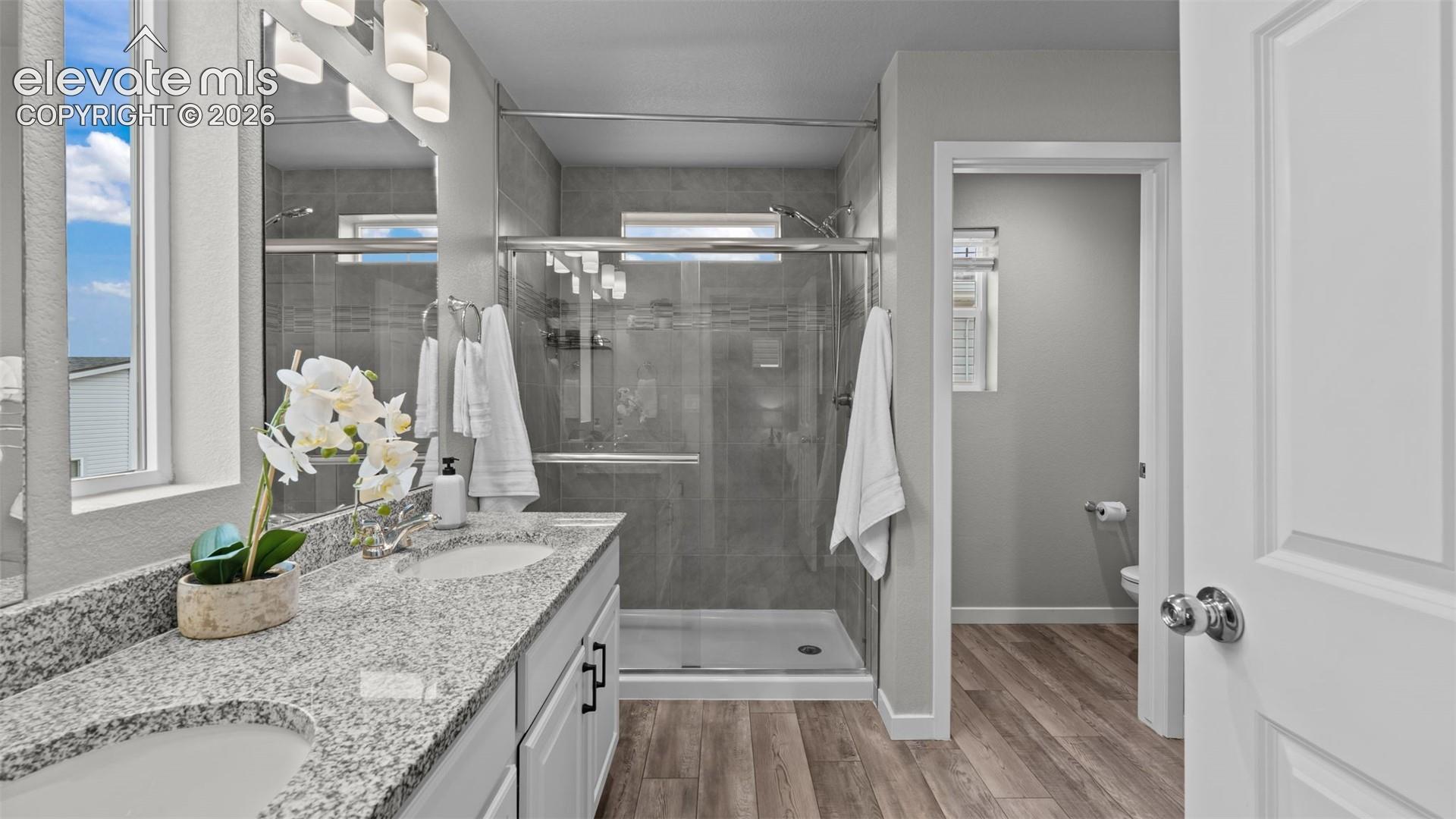 Dual-sink vanity with stone-style countertops, walk-in shower featuring glass enclosure and tile surround, and wood-finish flooring