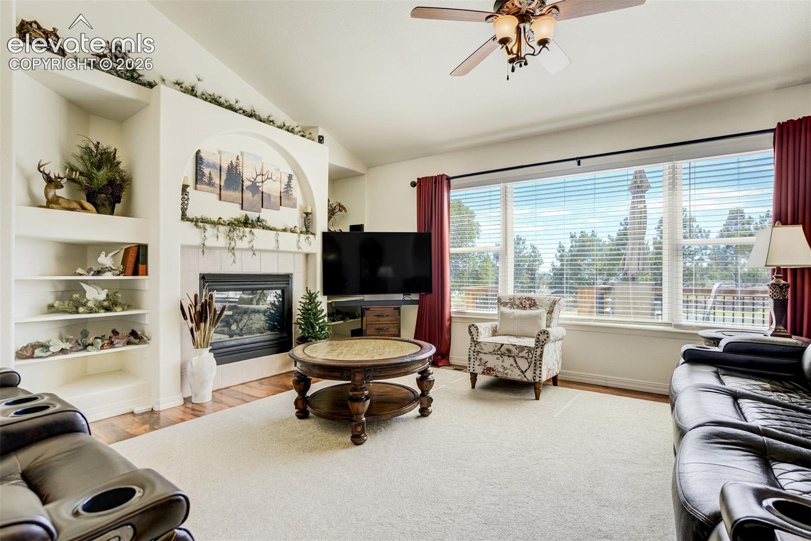 Living area featuring a vaulted ceiling, built-in shelving, a fireplace with an arched hearth, and expansive windows