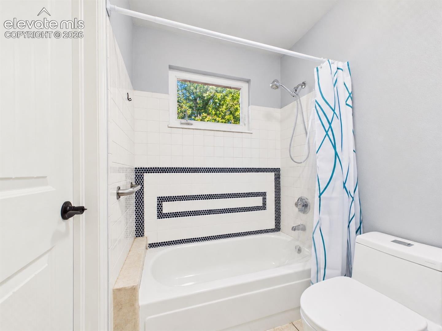 Tub and shower surround features white tile accented by a geometric dark mosaic design, plus a window that provides natural light.