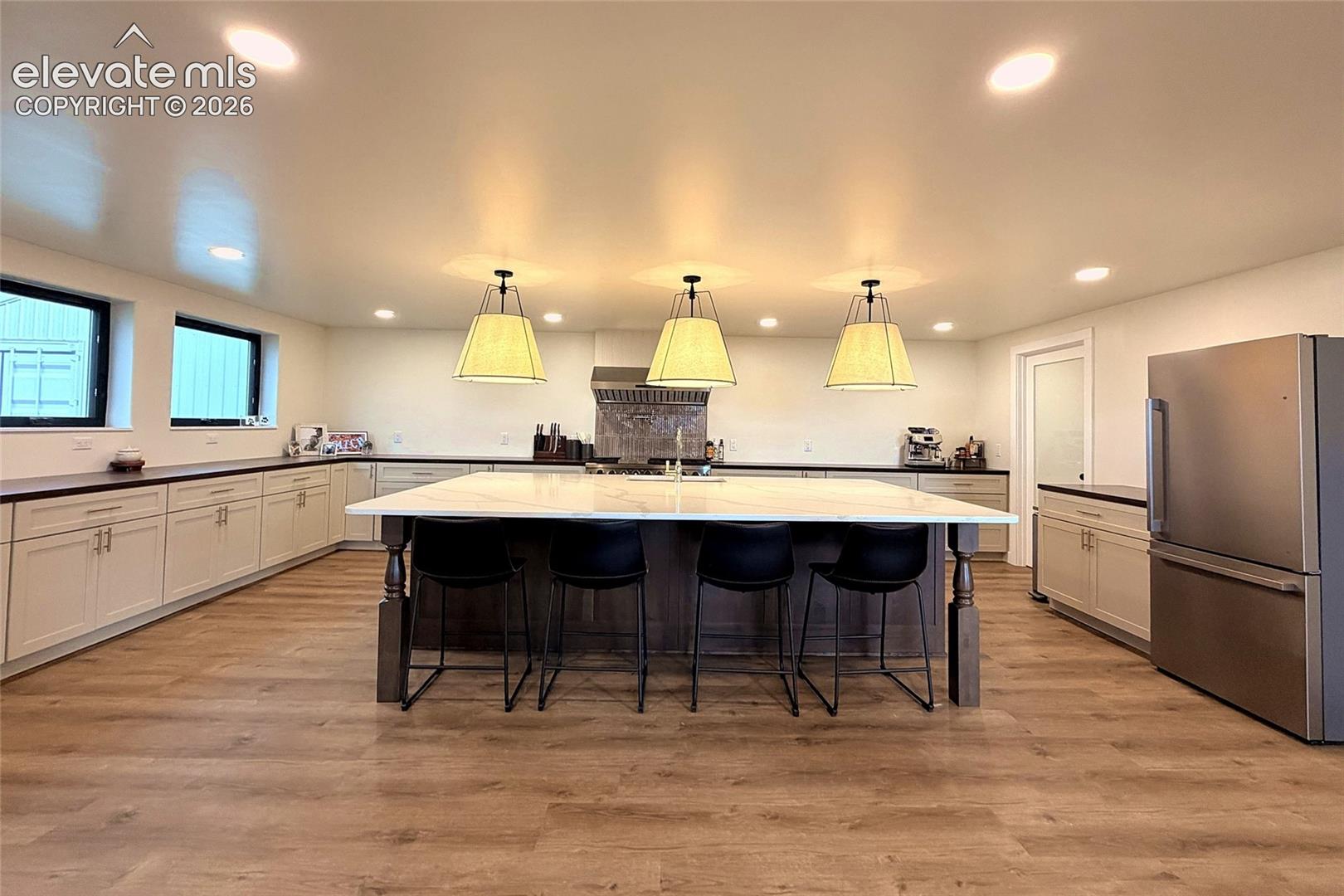 Spacious kitchen featuring an expansive center island with stone-surface countertops, custom shaker-style cabinetry, stainless steel appliances, wood-finish flooring, and trio pendant lighting