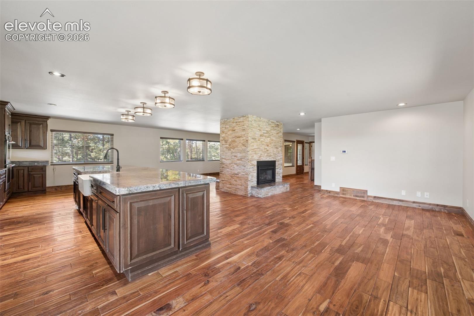 Open-concept interior featuring wood-finish flooring, a freestanding floor-to-ceiling stone fireplace, a kitchen island with stone surfacing, apron-front sink, and custom cabinetry