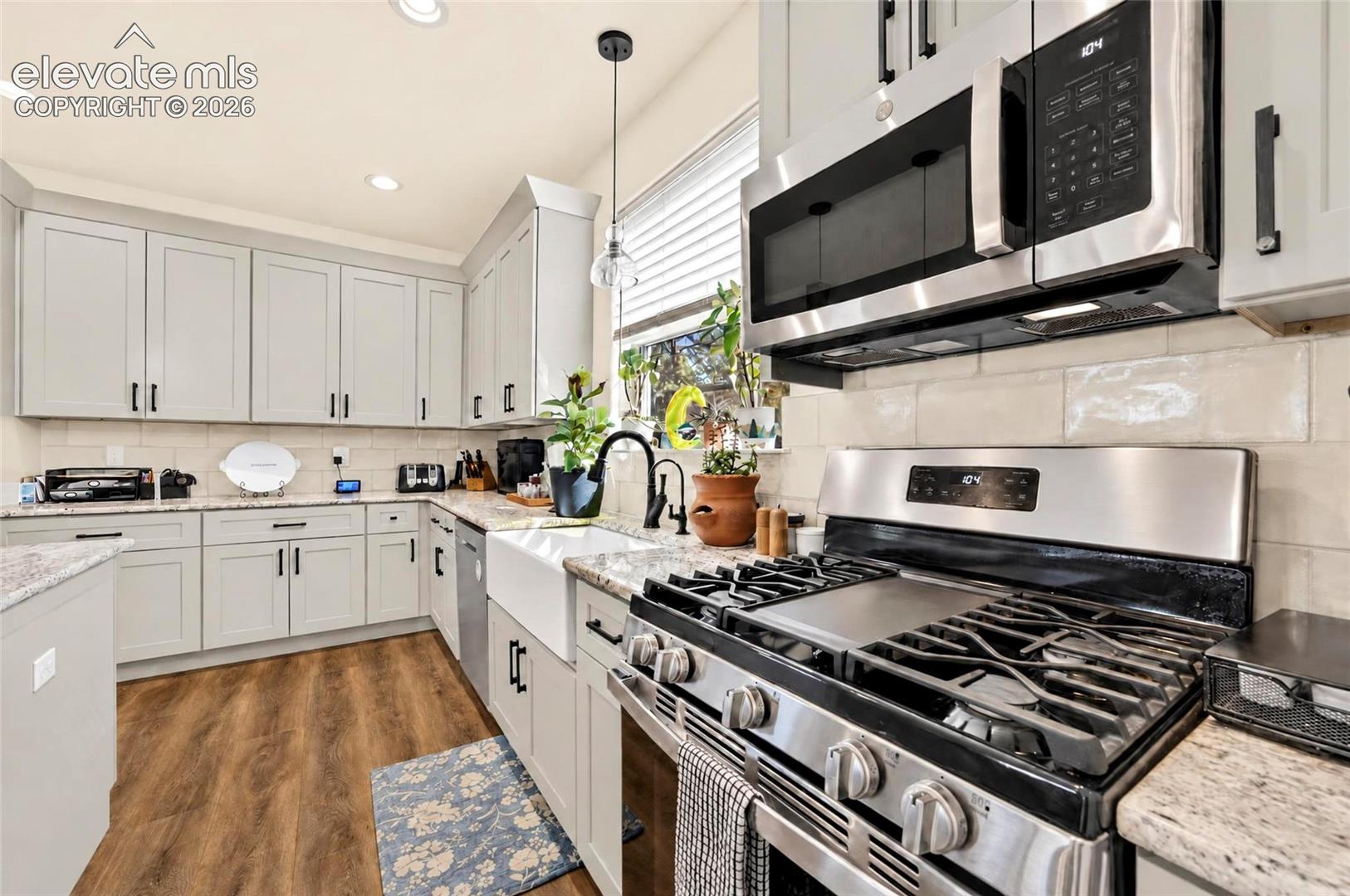 Kitchen featuring light grey cabinetry with matte black hardware, stone-finish countertops, subway tile backsplash, white farmhouse-style apron sink with matte black faucet, and wood-finish flooring