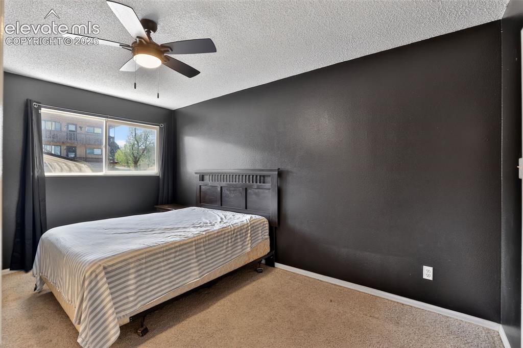 Bedroom featuring neutral-toned carpeting, dark accent walls, a window with exterior building and tree scenery, white baseboards, and a ceiling fan with integrated lighting