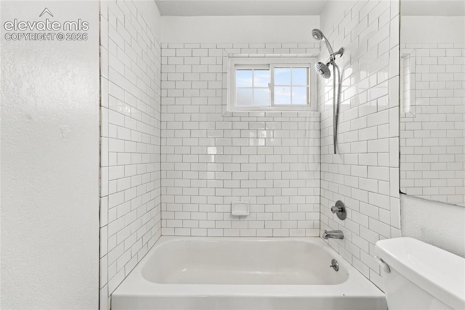 Bathroom featuring white subway tile wall surround, standard bathtub, chrome handheld shower head, slider window, and chrome faucet fixtures