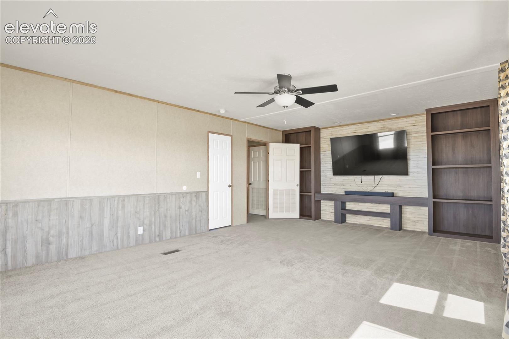 Living area featuring neutral carpeting, dual wood-finish built-in shelving units, a horizontal plank accent wall, and a ceiling-mounted light fixture with fan blades