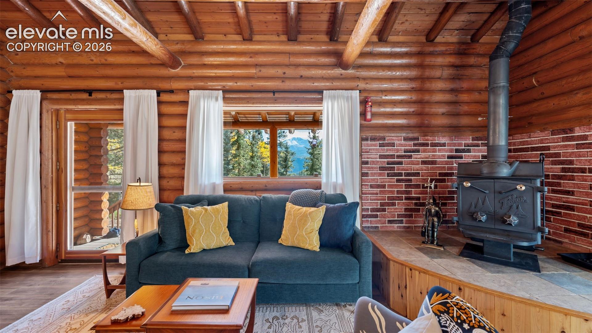 Log walls, exposed wood ceiling beams, wood-finish flooring, metal wood-burning stove with brick surround, and stone-tiled hearth