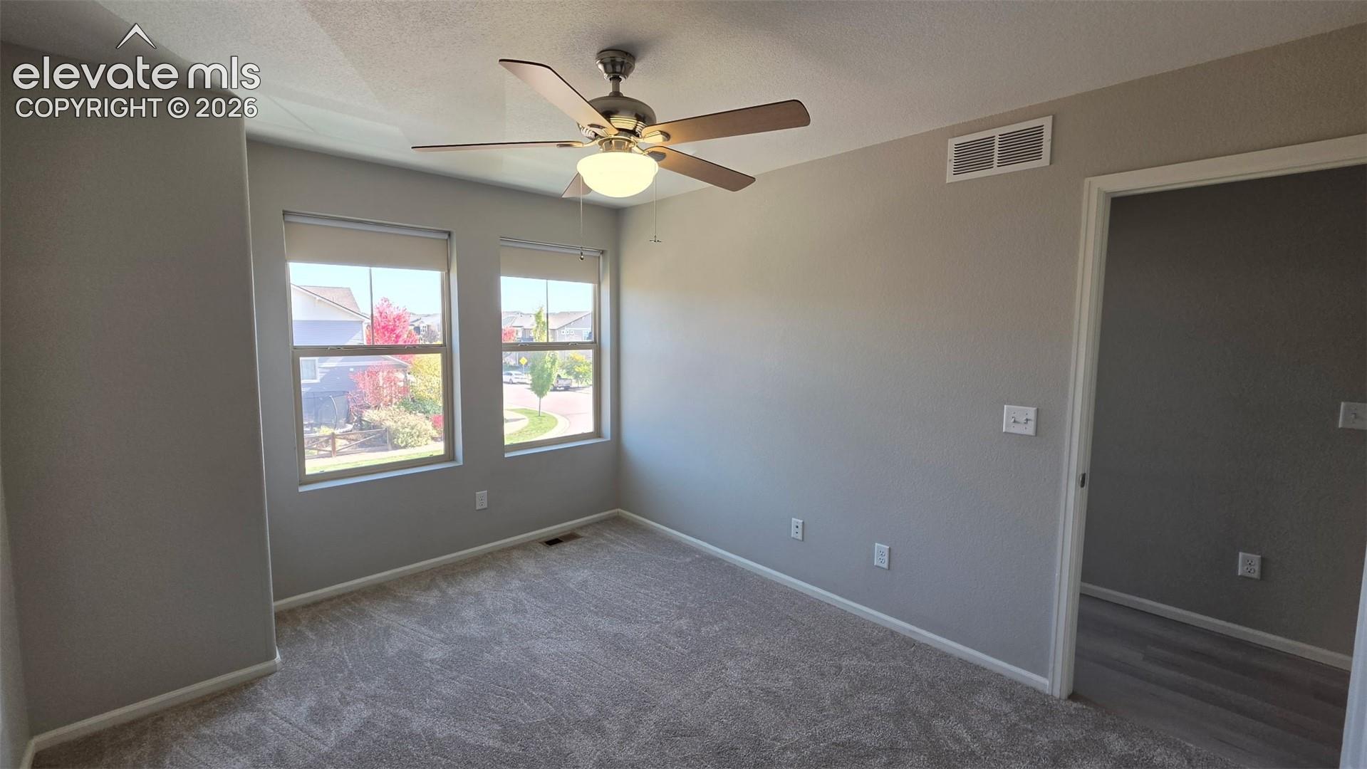 Neutral-toned interior room featuring wall-to-wall carpeting, dual windows with roller shades, and a ceiling-mounted light fixture with a multi-blade fan
