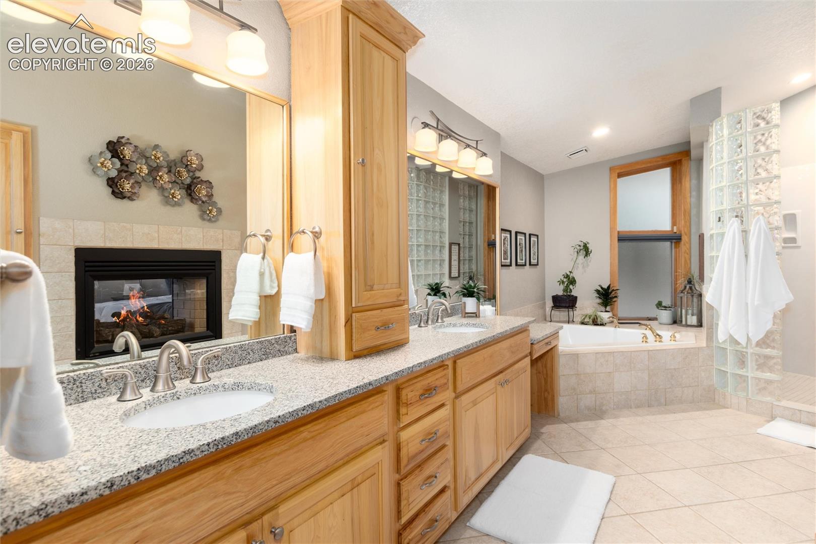Spacious bathroom featuring a dual vanity with granite countertops, built-in cabinetry, an oversized soaking tub, a glass block shower enclosure, and a fireplace