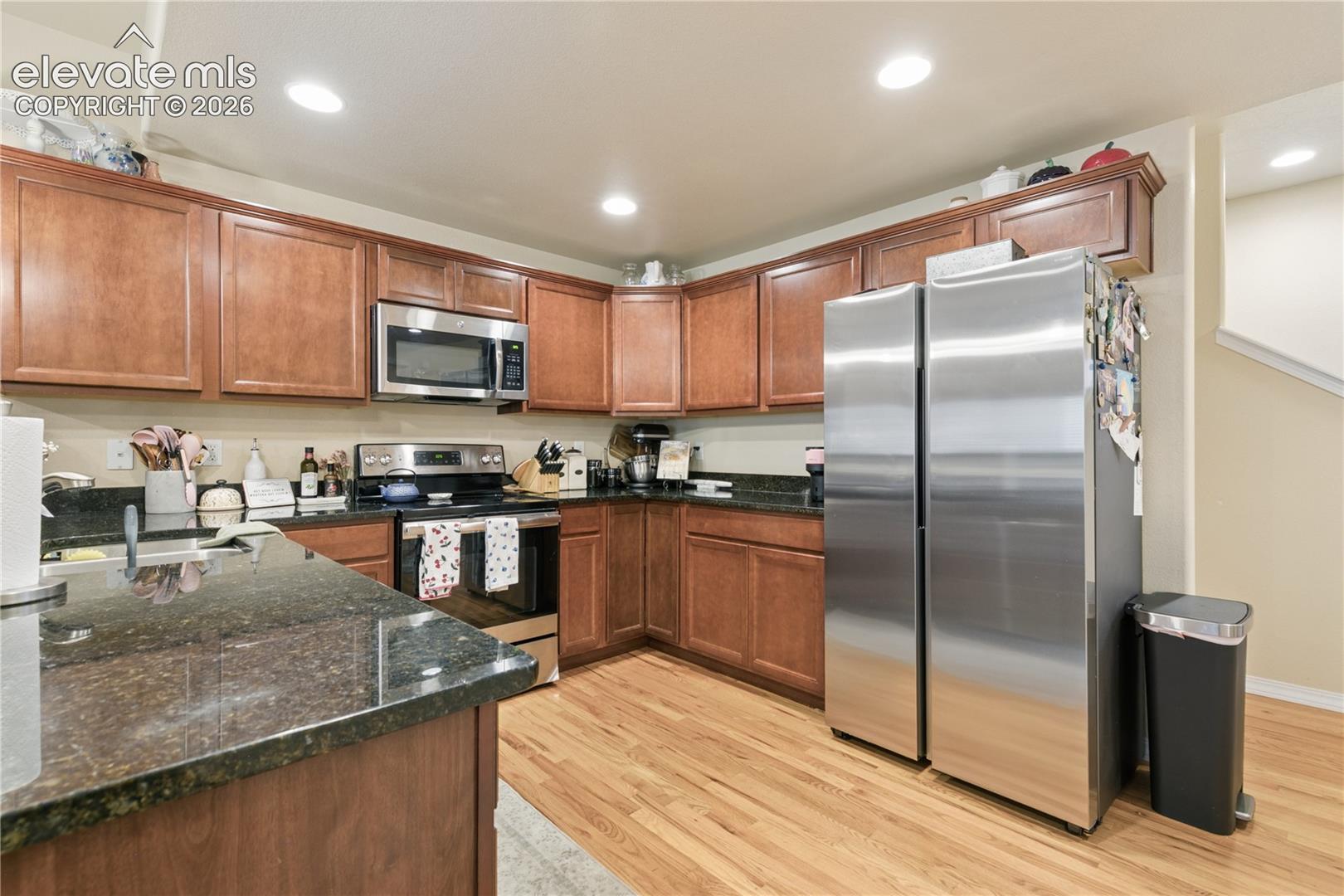 Kitchen featuring wood-finish flooring, dark stone countertops, wood cabinetry, and stainless steel appliances including a side-by-side refrigerator, microwave, and range