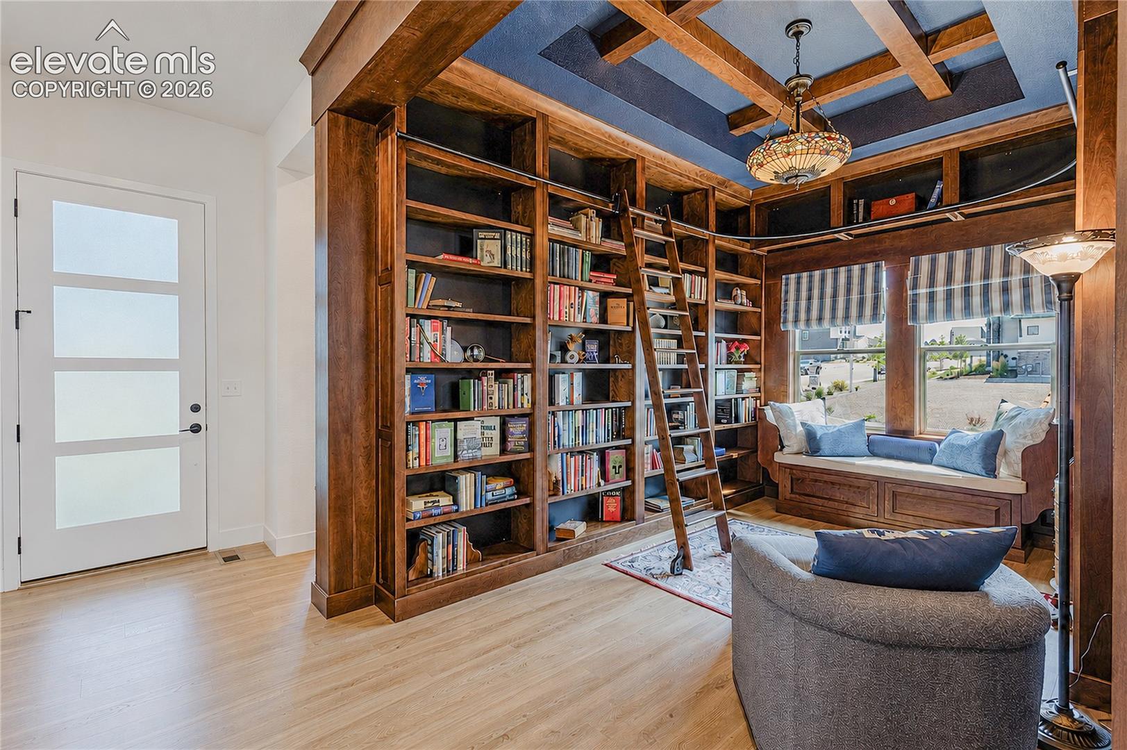 Custom built-in bookcases with a rolling ladder, coffered ceiling with a central pendant fixture, and a built-in window seat