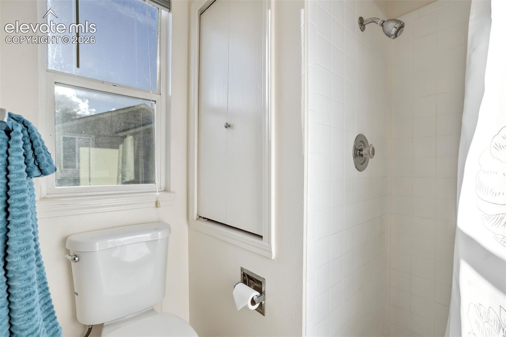 Shower surround featuring white square ceramic tile, chrome shower hardware, built-in wall cabinet, and operable window with natural light