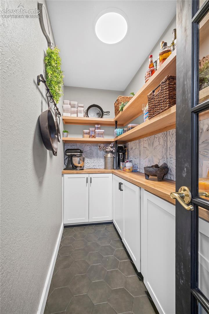 Pantry featuring custom wood shelving, white cabinetry with wood-finish countertops, decorative patterned tile backsplash, dark hexagonal floor tiling, and a black-framed glass panel door