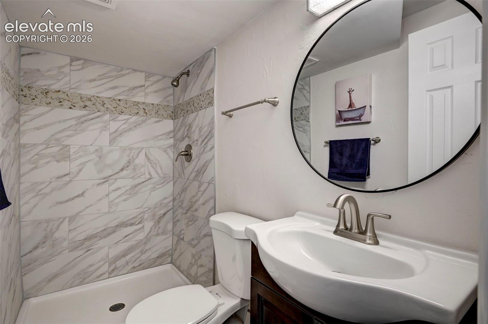 Full bathroom featuring a shower stall with stone-patterned tile surround, decorative mosaic accent banding, oval wall-mounted mirror, and an integrated vanity sink with metallic hardware