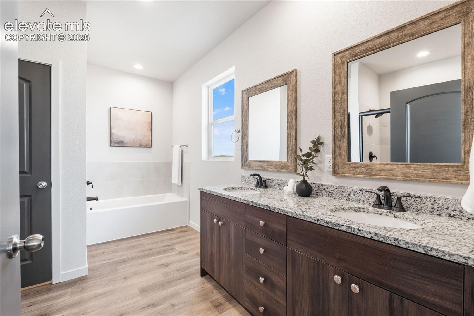 Double vanity with granite countertops and undermount sinks, wood-finish flooring, recessed lighting, and a built-in tub with tile surround