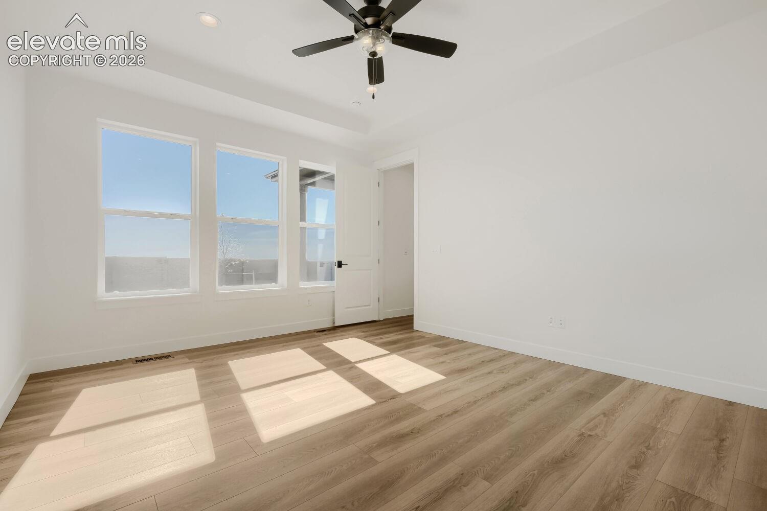 Bright, light-filled bedroom featuring expansive windows with natural illumination, wood-finish flooring throughout, and a ceiling fan with integrated lighting