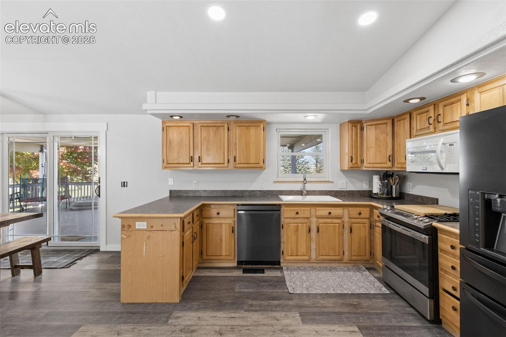 Kitchen featuring wood cabinetry, dark-finish countertops, recessed lighting, integrated black appliances, and wood-finish flooring