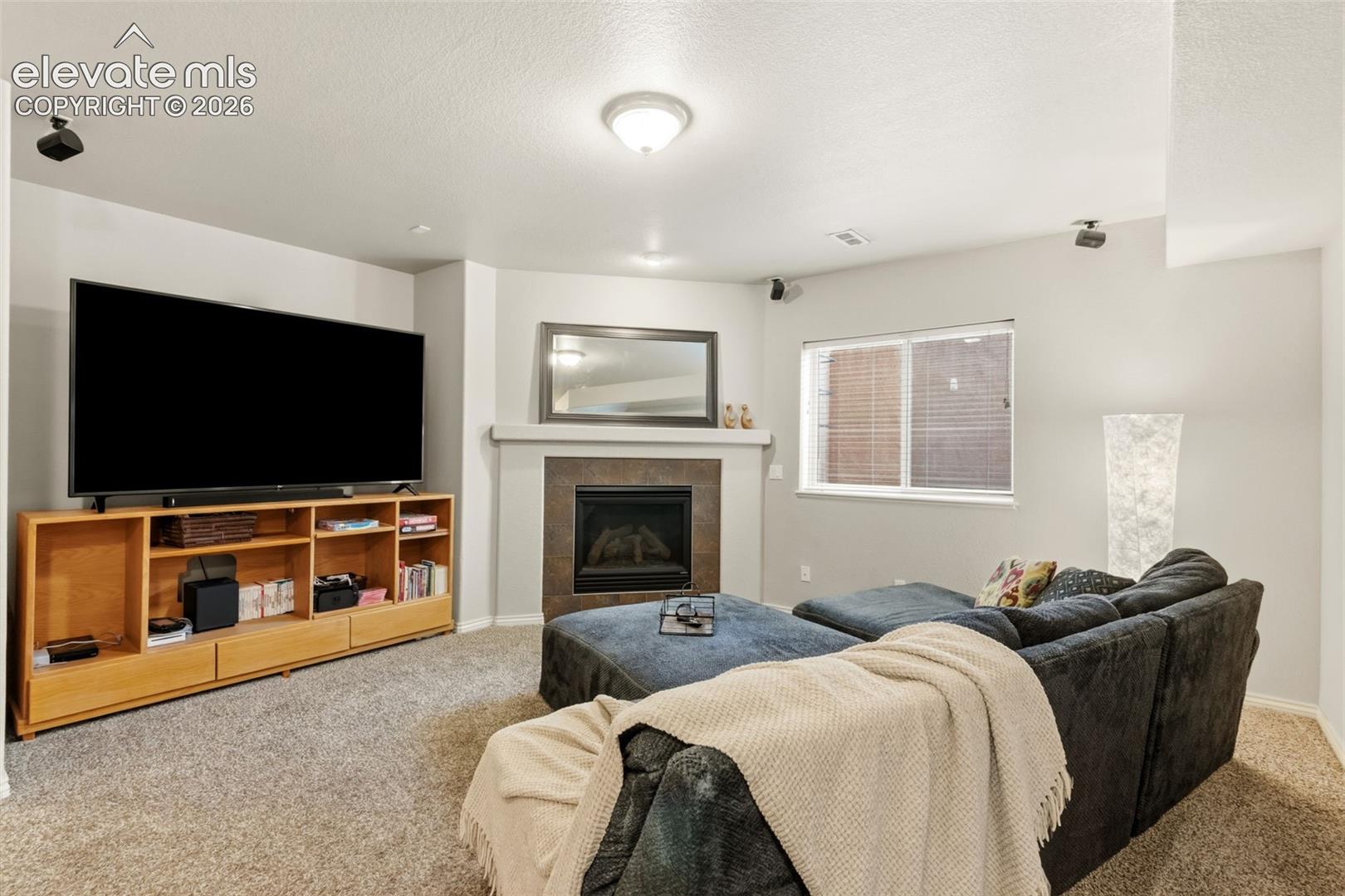 Carpeted living space featuring a fireplace with stone surround and mantle, a window with blinds, and built-in ceiling speakers