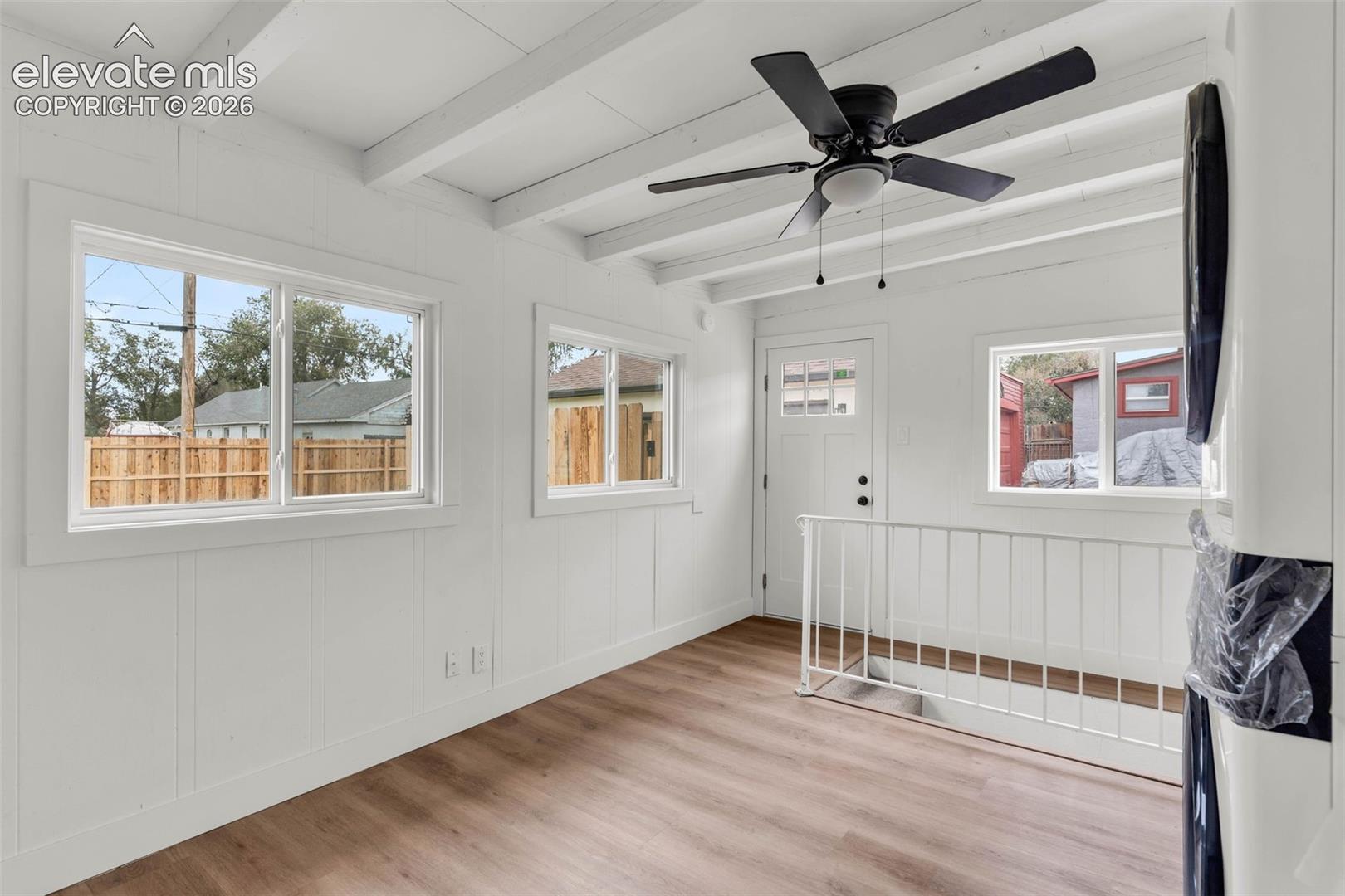 White wood-finish flooring and beamed ceiling featuring a black matte finish ceiling fan, three double-pane sliding windows, and white metal railing