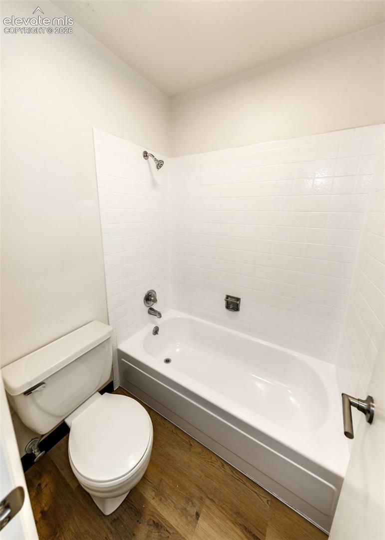 Bathroom featuring wood-finish flooring, a white porcelain toilet, and a bathtub with white tile surround, chrome faucet, and integrated shower head