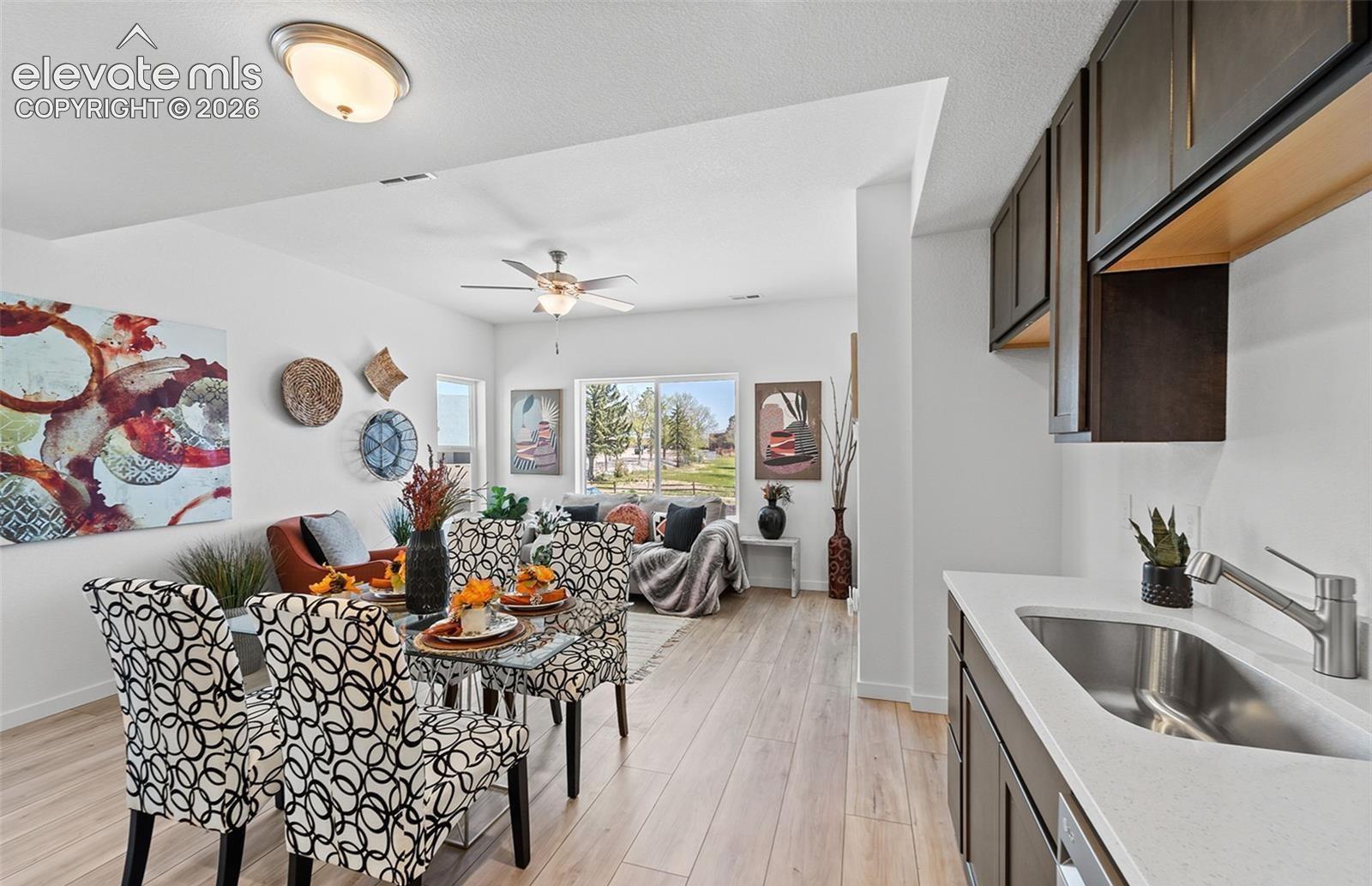 Open-concept area featuring wood-finish flooring, dark cabinetry, light-toned stone countertops, an integrated stainless steel sink with a high-arch faucet, and a ceiling fan with integrated lighting