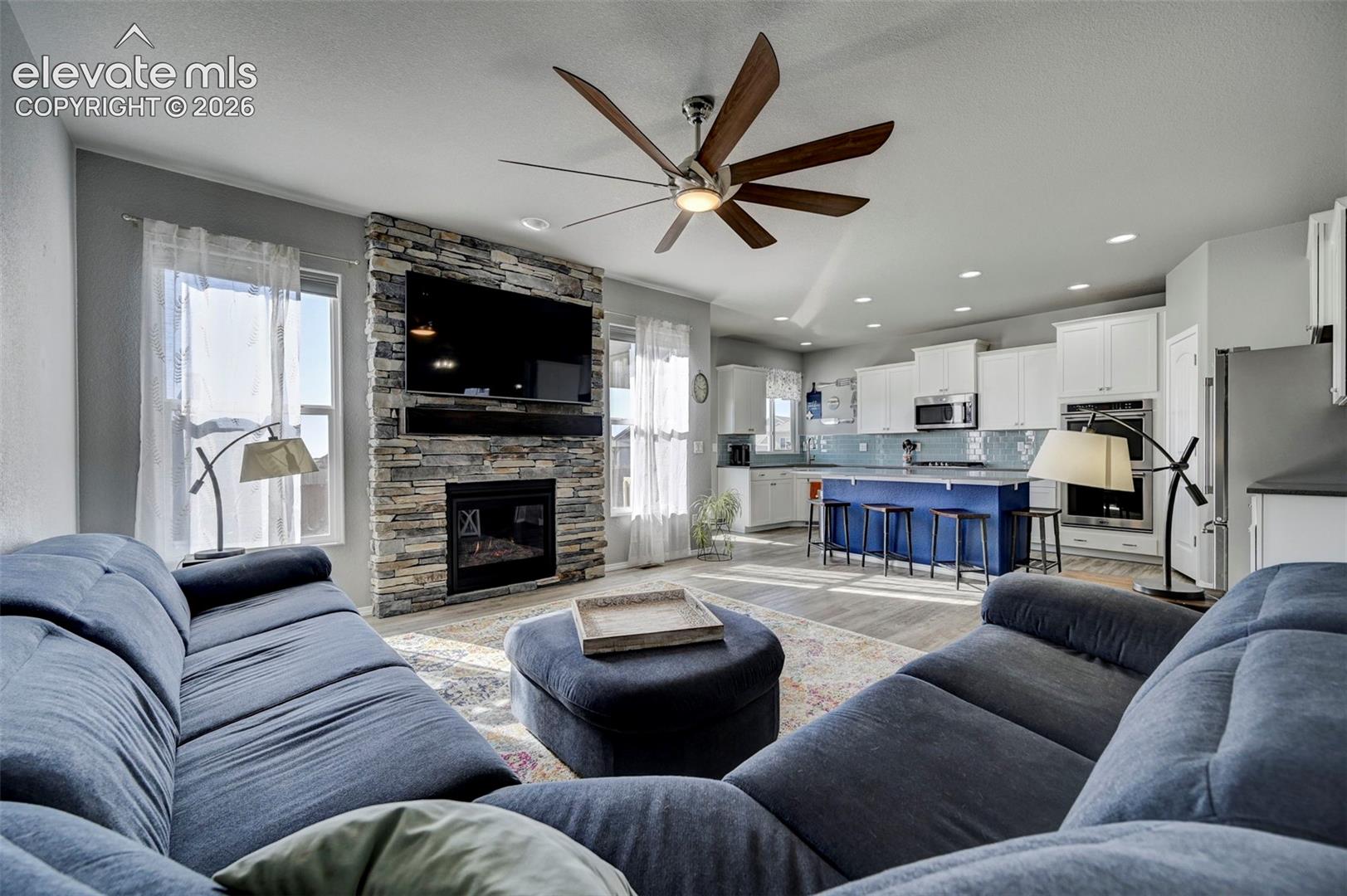 Open-concept living area featuring a stacked stone fireplace with a built-in mantel, a ceiling fan with integrated lighting, recessed lighting, and wood-finish flooring