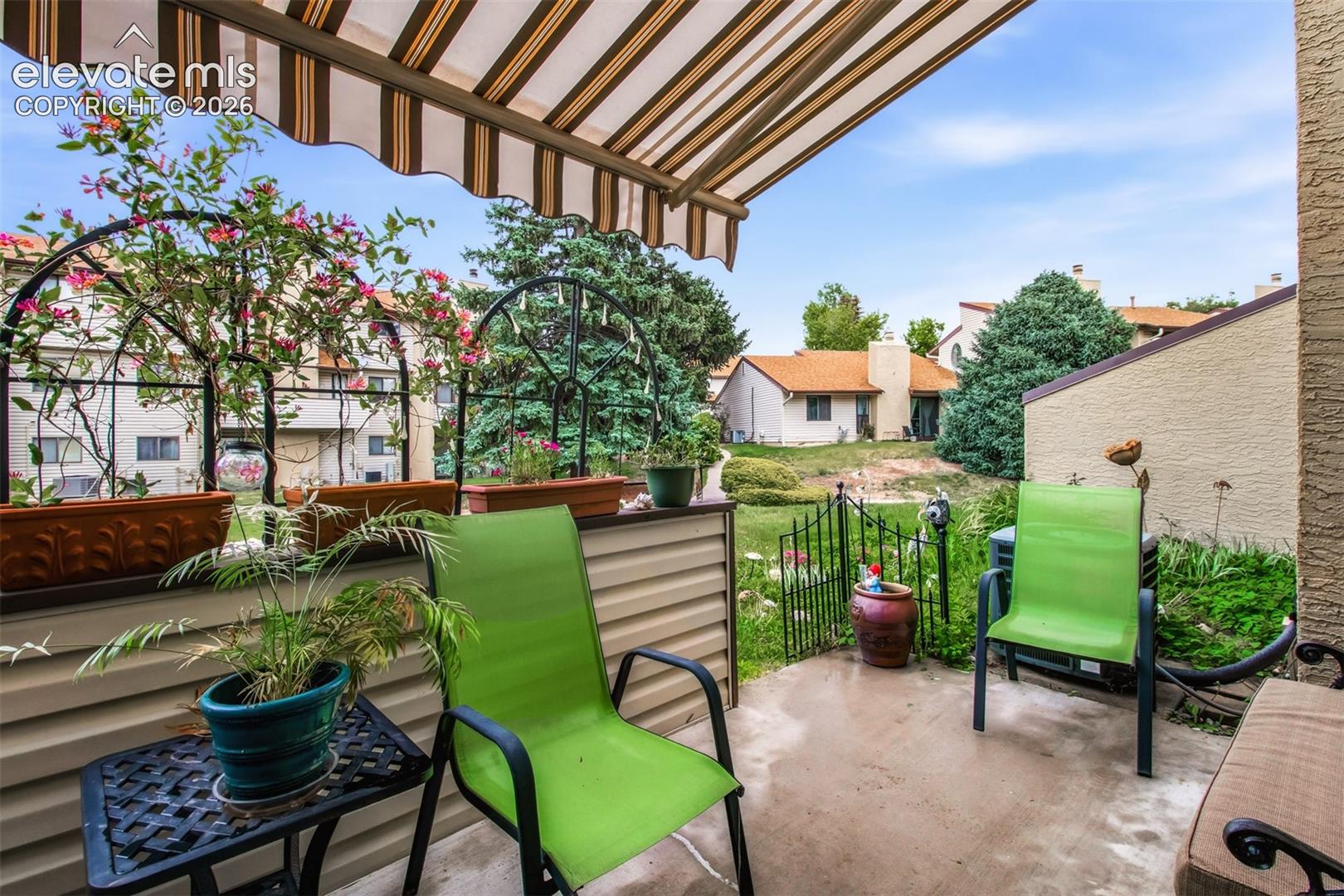 Concrete patio featuring a retractable striped awning, neutral siding wall, and metal garden gate
