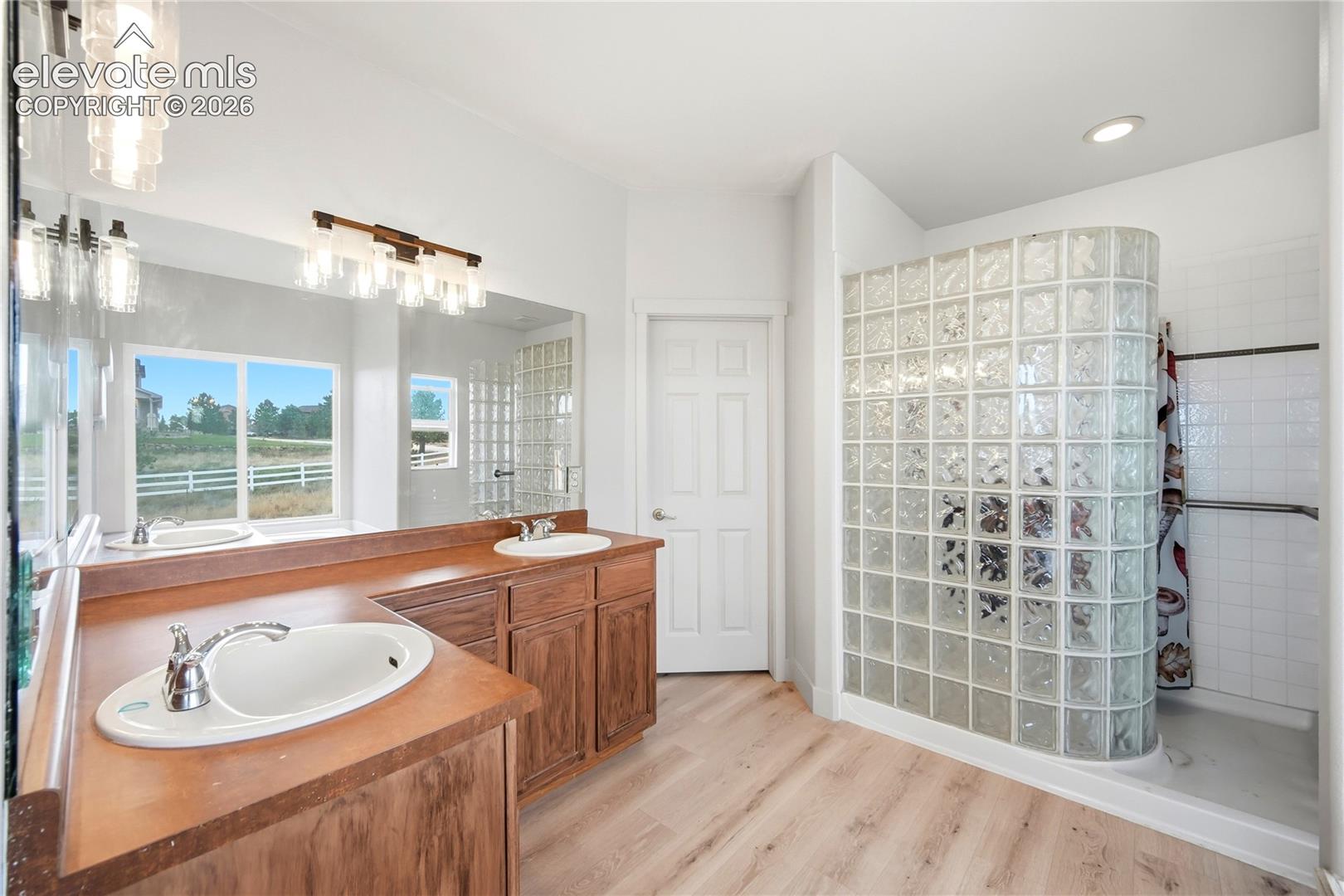 Bathroom featuring wood-finish flooring, a dual-sink vanity with a wood countertop, a full-width wall mirror, and a curved glass block walk-in shower