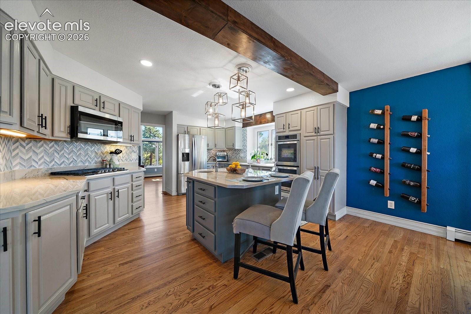 Kitchen featuring wood-finish flooring, a central island with a stone-surface countertop, herringbone-patterned tile backsplash, stainless steel built-in appliances, and a rustic timber ceiling beam