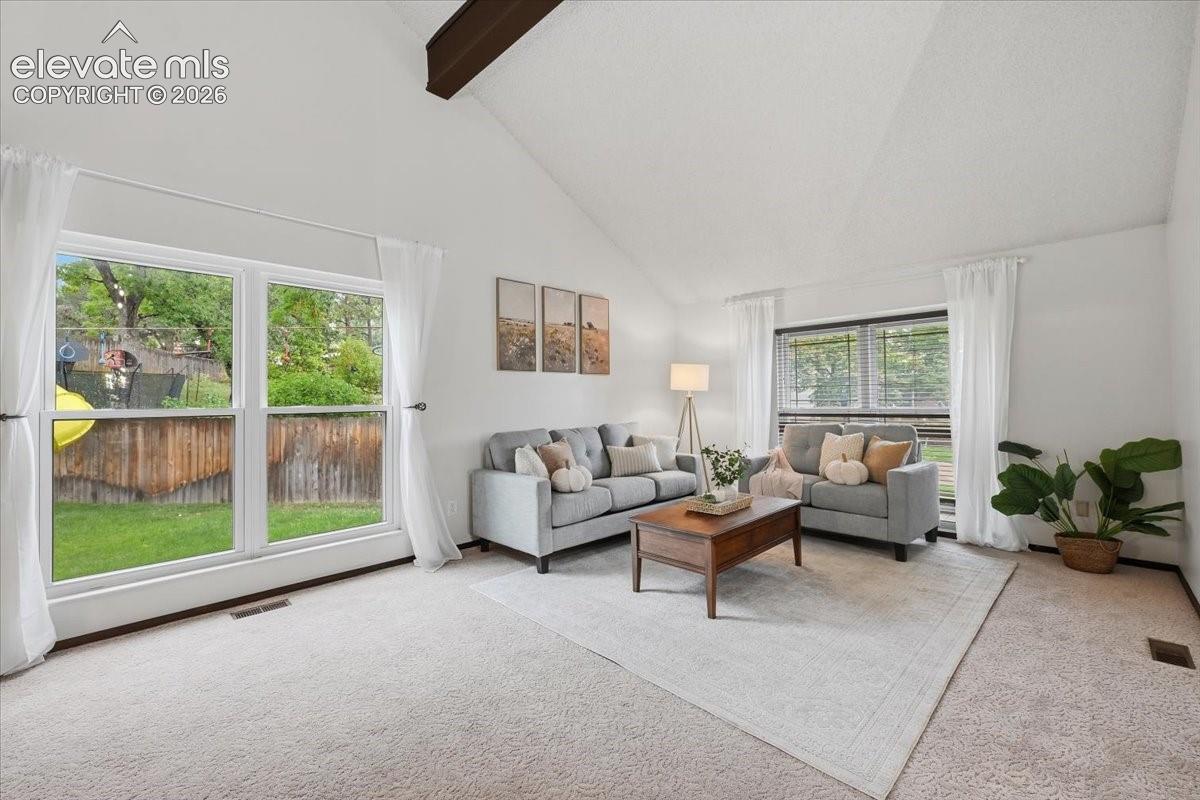 Bright, light-filled living room featuring vaulted ceilings with exposed wood beams, expansive glass windows overlooking manicured grounds