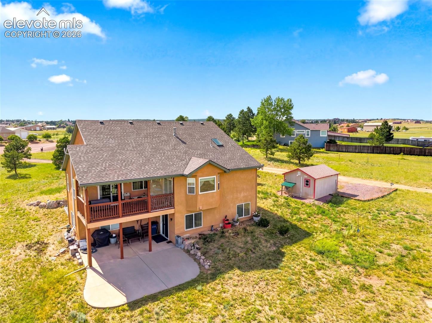 Spacious two-story residence with warm exterior finish and deck, situated on expansive grounds with mature landscaping, detached outbuildings, and open acreage